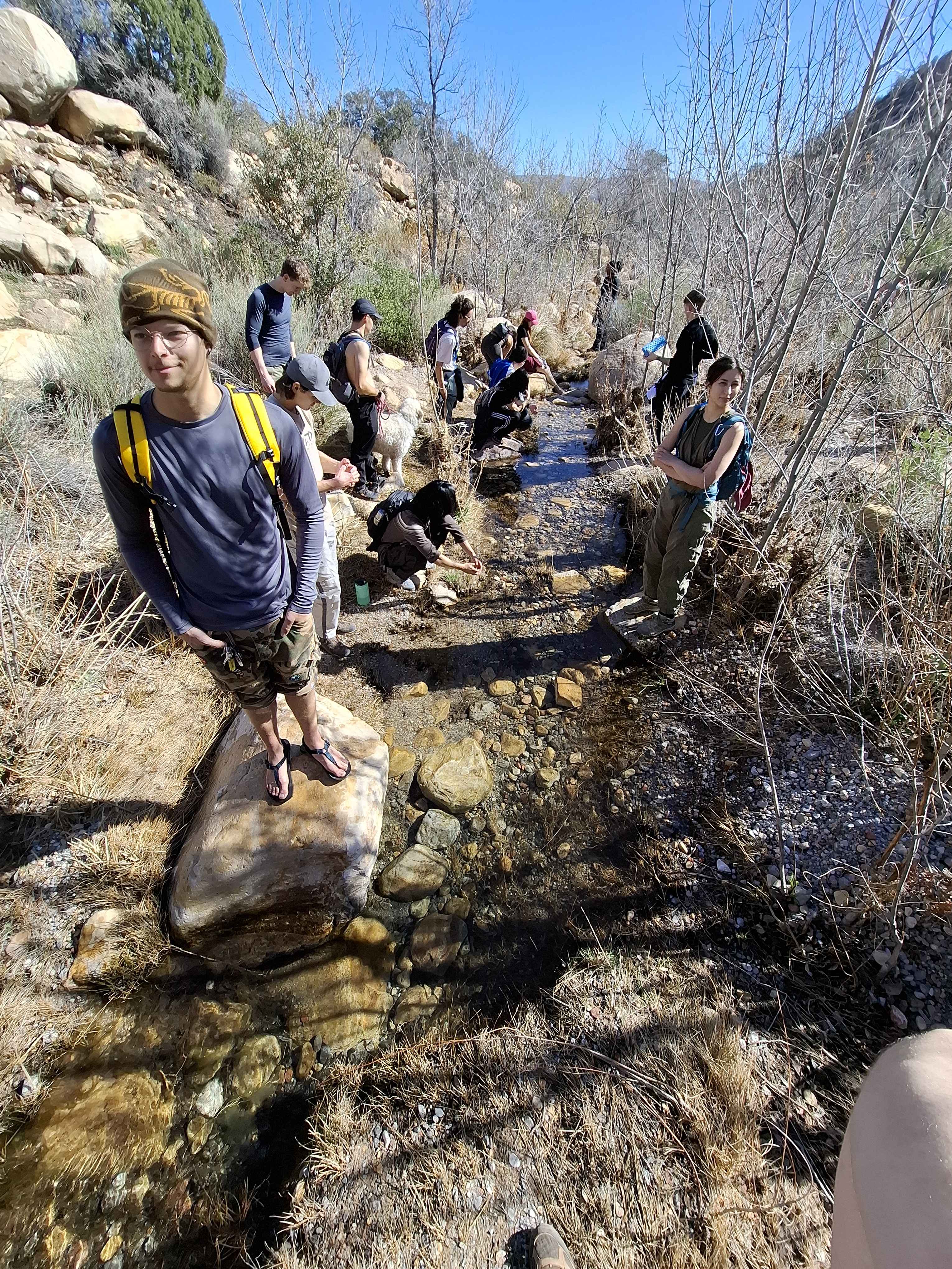 Mountain Club hikers walk along a shallow creek through a rocky, leafless canyon.