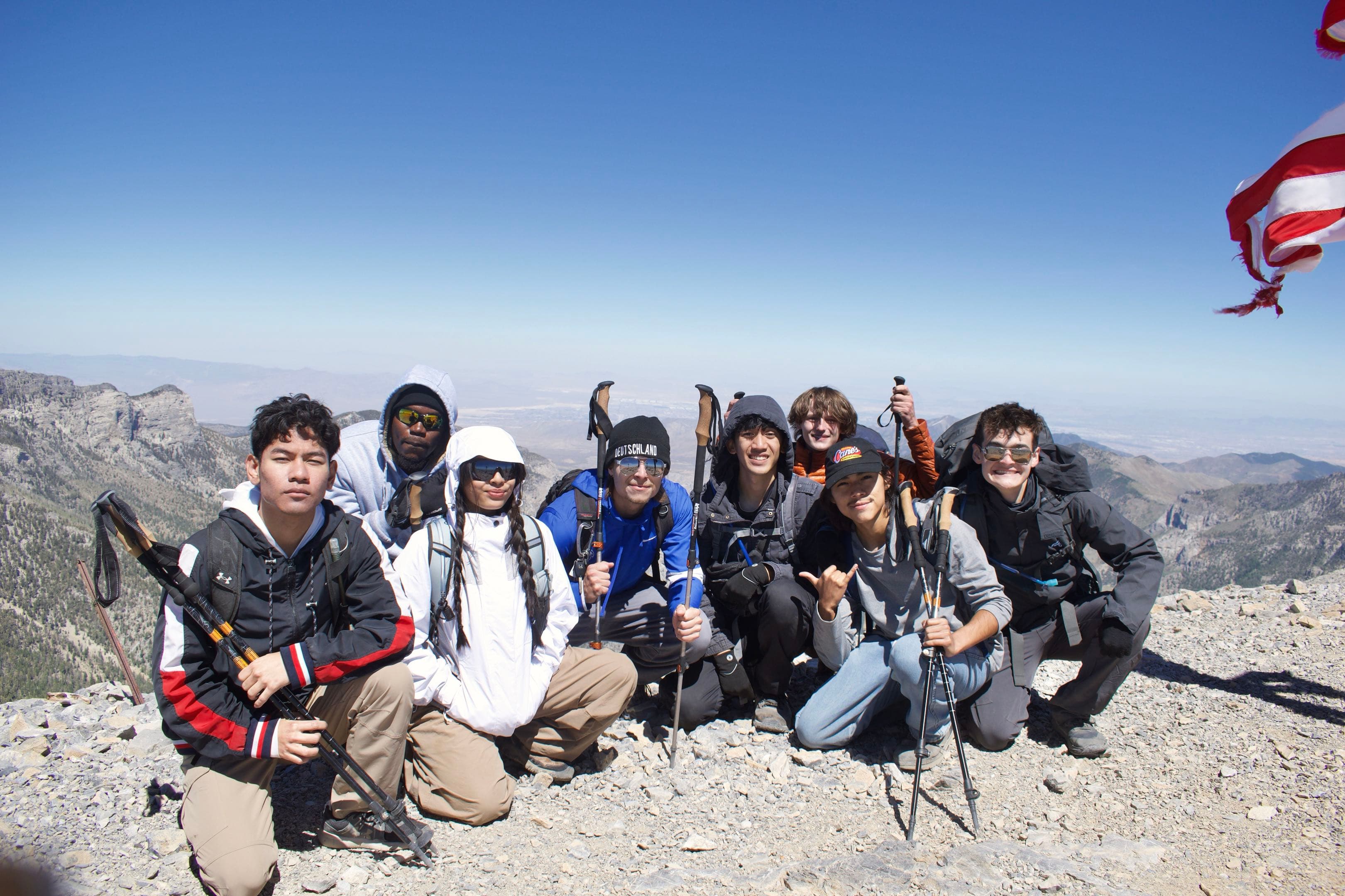 A group of Mountain Club hikers kneel together at a rocky desert summit beneath a blue sky.