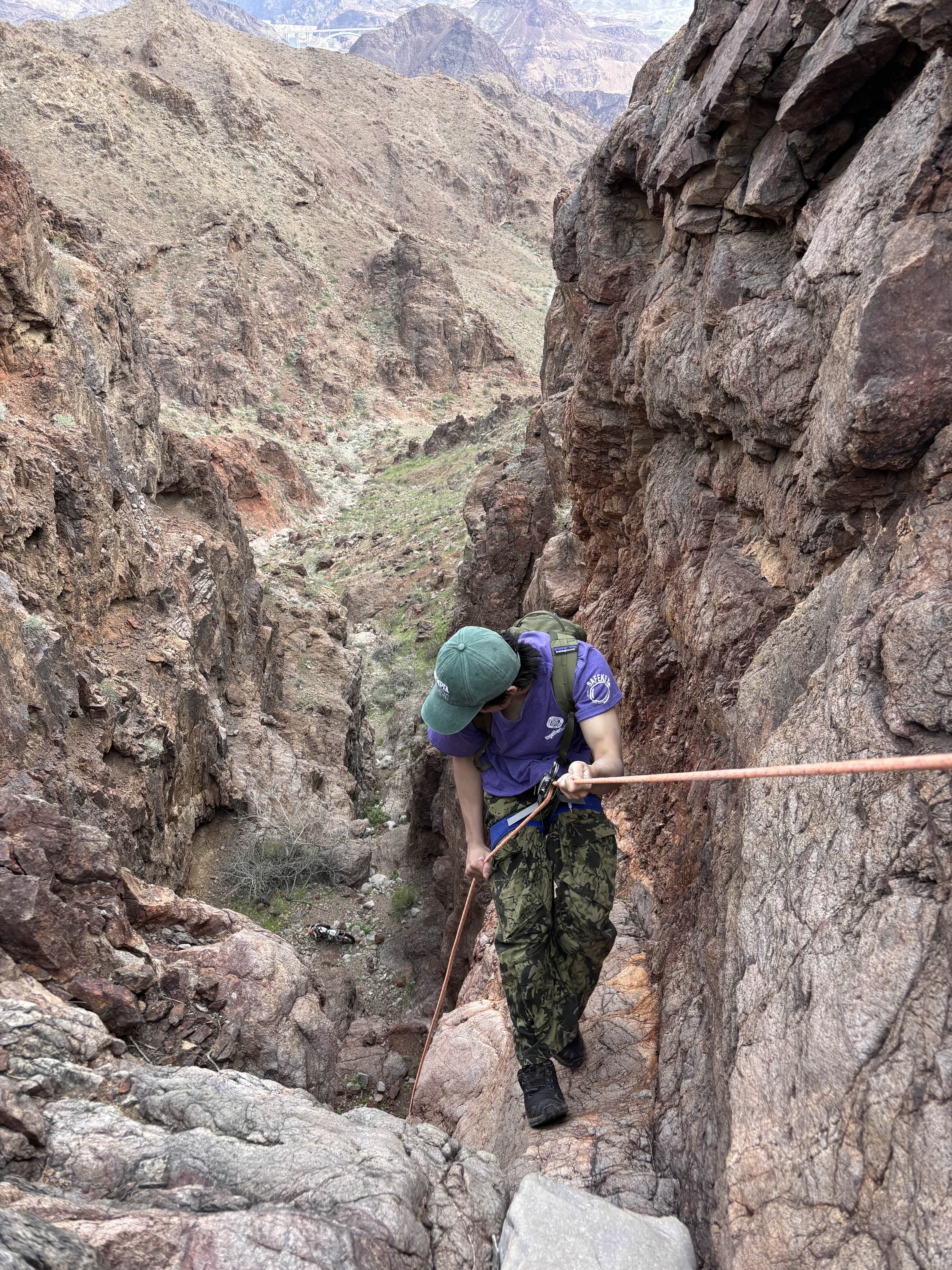 A helmeted climber rappels between steep canyon walls while a club member watches from below.