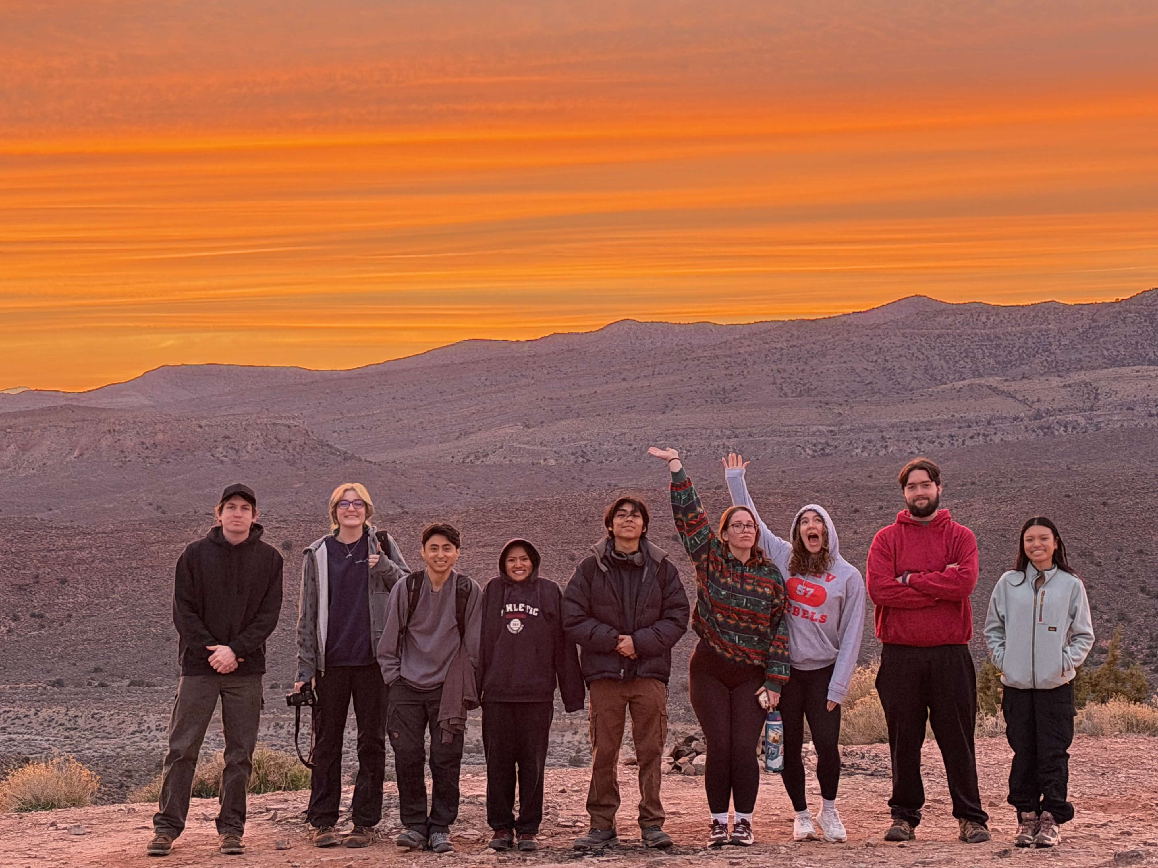 Club members stand together in the desert as orange sunset clouds stretch above the mountains.