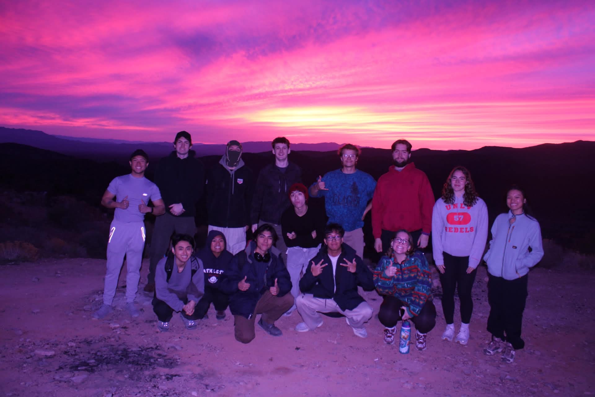 A large group of club members pose together beneath a bright pink desert sunset.