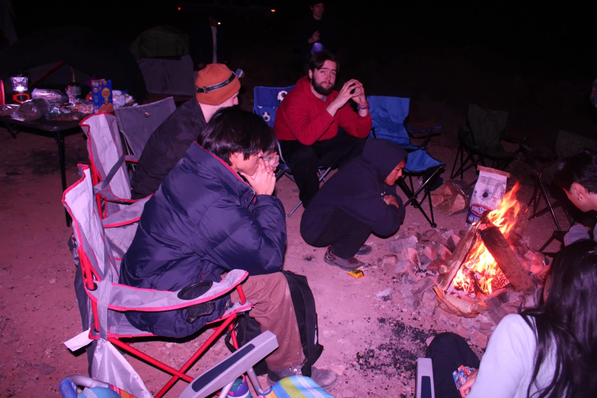 Club members gather in camp chairs around a small nighttime campfire.