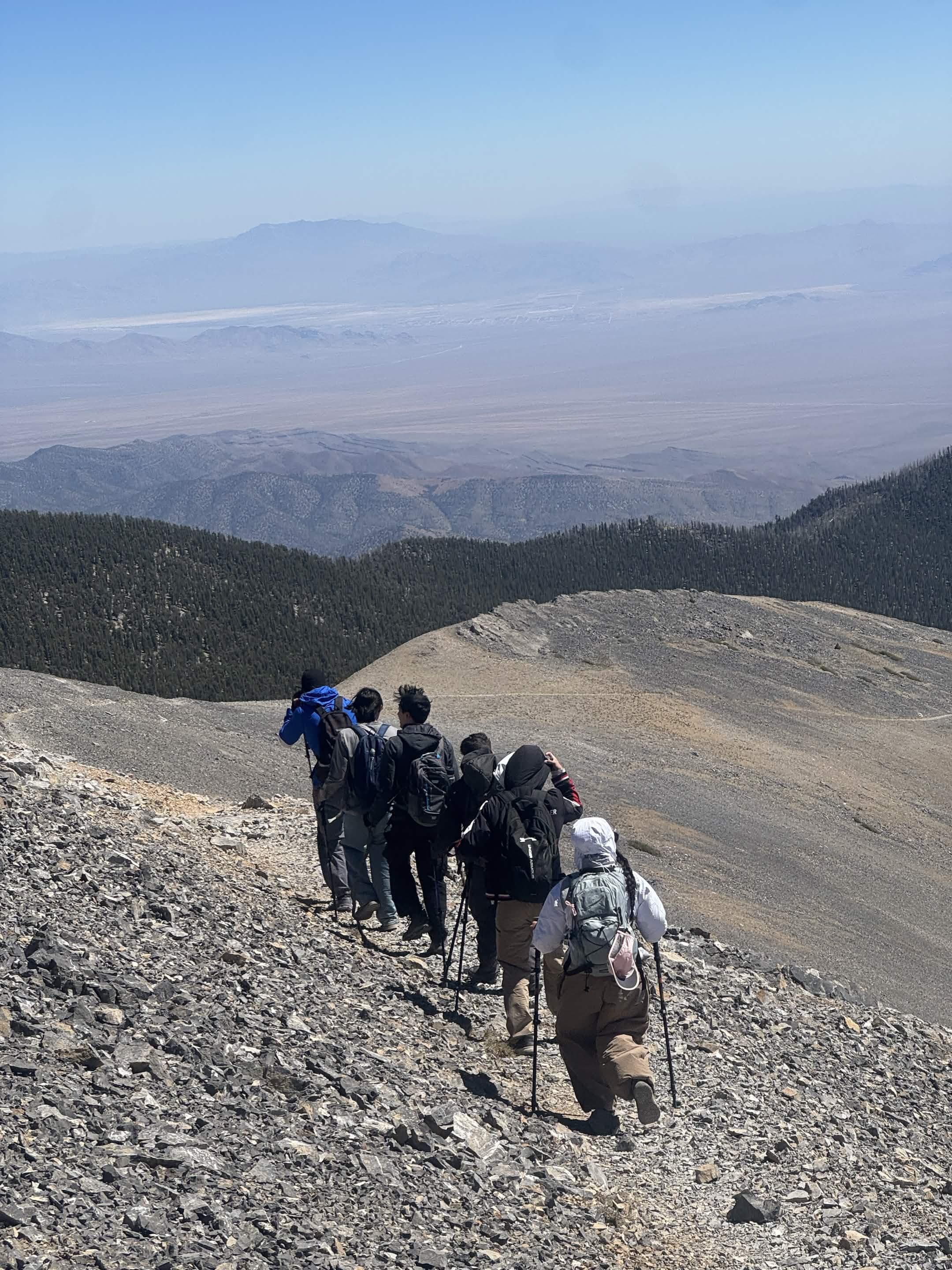 A line of hikers climbs a pale rocky mountain slope above the desert valley.
