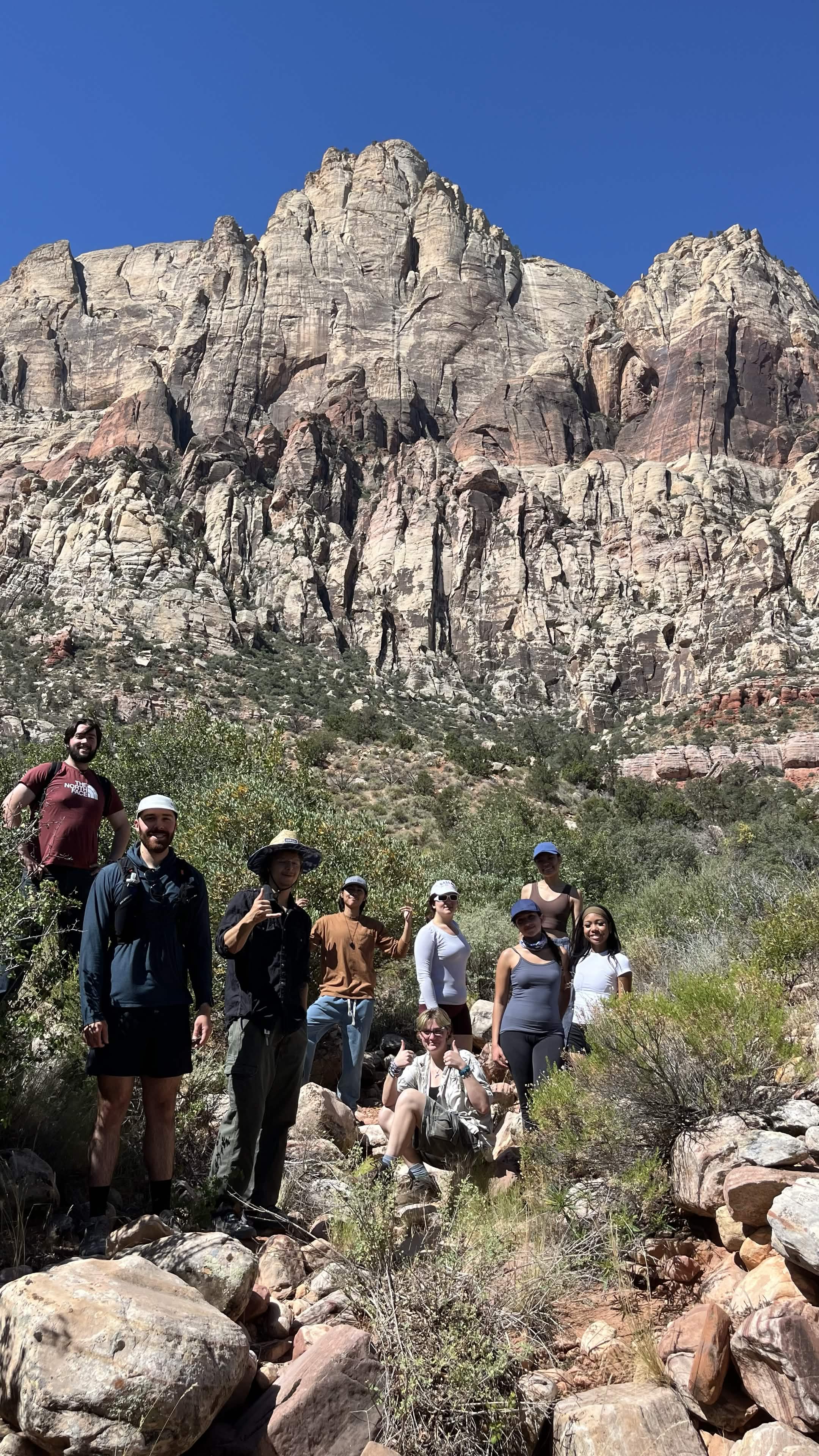 A group of hikers pauses beneath tall pale cliffs and a clear blue sky.