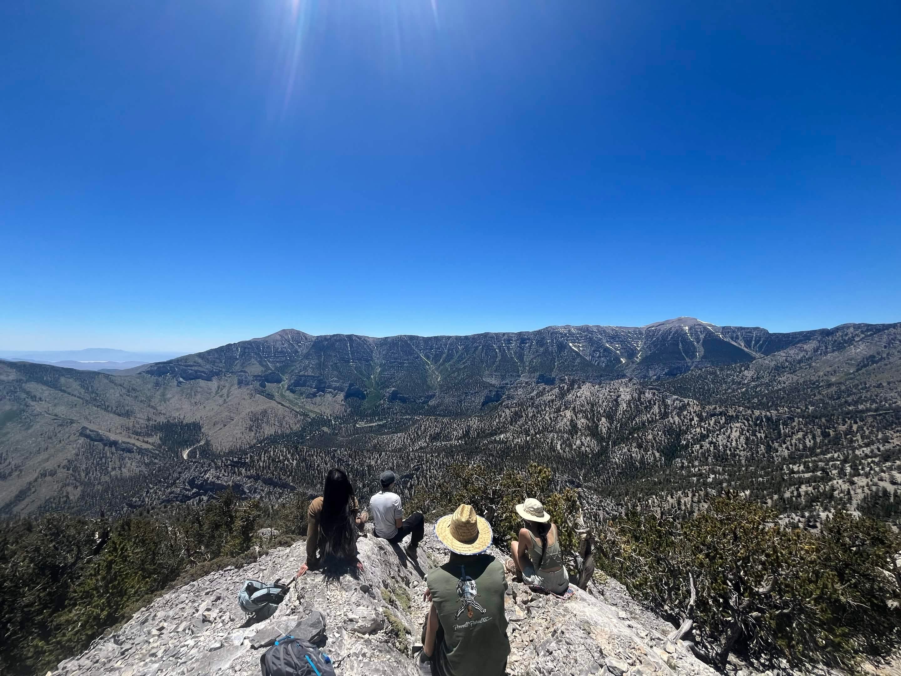 Hikers sit at a rocky overlook above a wide forested mountain landscape.