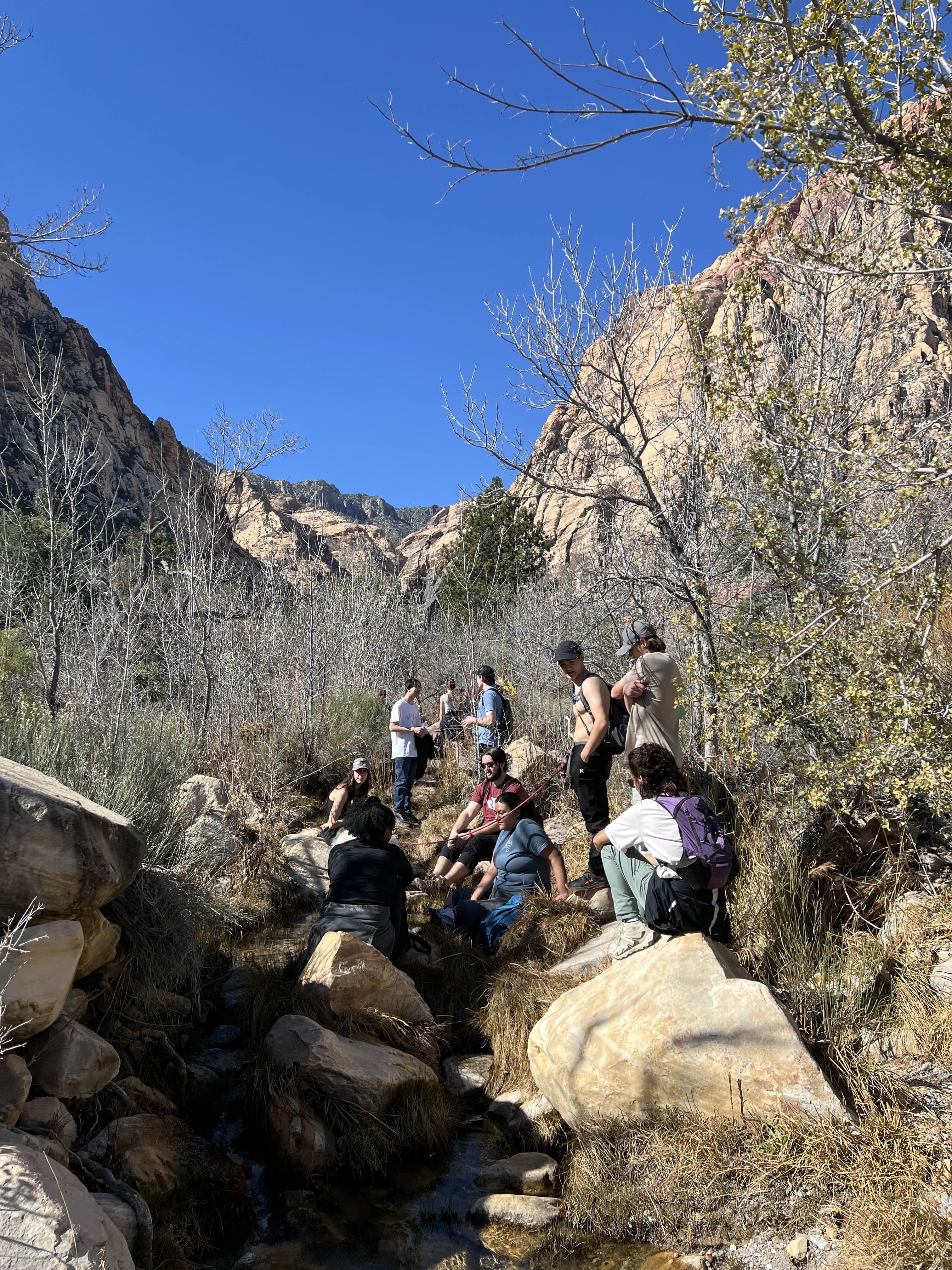 A group of hikers sits together on large rocks at the base of a steep canyon wall.